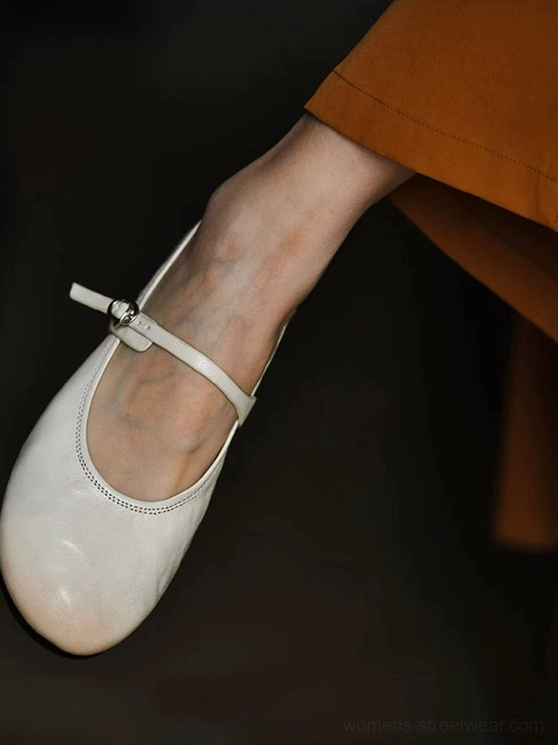 Close side view of an ivory leather Mary Jane flat on foot with a caramel skirt, highlighting the narrow ankle strap and polished silver buckle