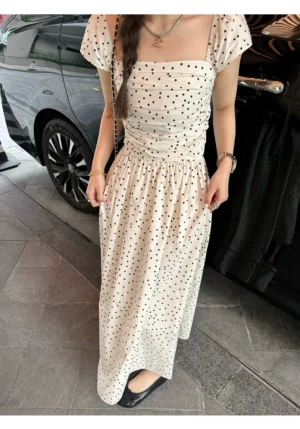 Ivory polka dot flutter sleeve maxi dress styled with layered necklaces beside a black sedan, showing the square neckline, ruched bodice, fitted waist, and flowing floor length skirt