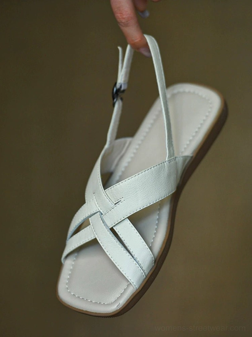 Ivory leather flat sandal held overhead, featuring crisscross front straps, square open toe, padded insole, and round buckle slingback strap
