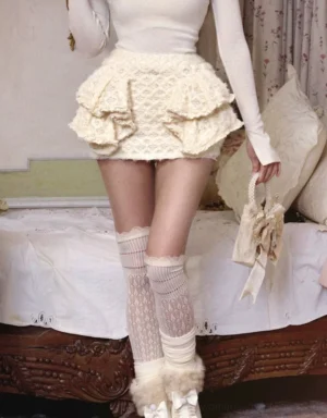 Rear low-angle view of ivory tiered ruffle knit mini skirt styled with cream knit top and fluffy leg warmers in a vintage bedroom