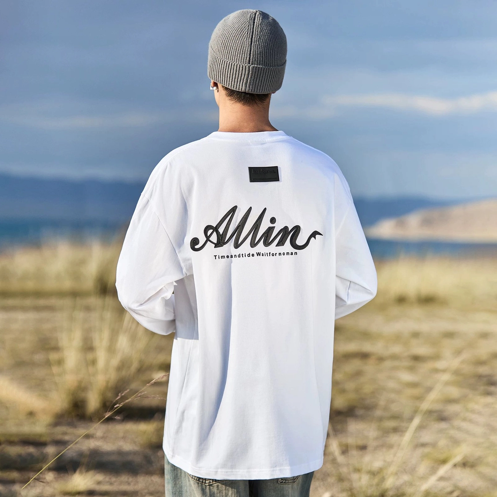 Ivory white alpine script oversized cotton long sleeve streetwear tee with bold black back logo beside a lakeside