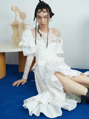 Ivory white asymmetric ruffle midi dress shown in a low angle seated studio look with puff sleeves, textured bodice, layered ruffled hem and romantic Korean streetwear styling