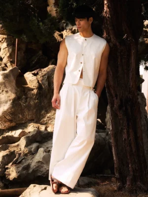 Male model wearing an ivory white minimal button front sleeveless streetwear vest with matching wide leg trousers in a rocky garden setting