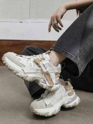 Women wearing khaki chunky patchwork mesh trail sneakers with utility straps and rugged platform soles styled with loose dark gray jeans in an industrial studio