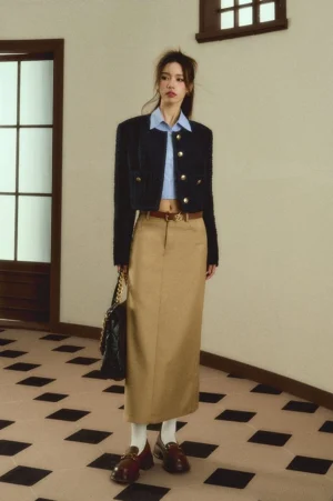 Full body front view of a woman wearing a khaki high waisted maxi skirt with a cropped tweed jacket and blue shirt in a vintage apartment