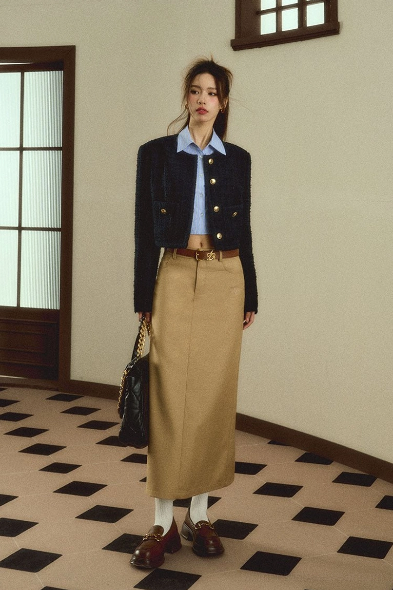 Full body front view of a woman wearing a khaki high waisted maxi skirt with a cropped tweed jacket and blue shirt in a vintage apartment