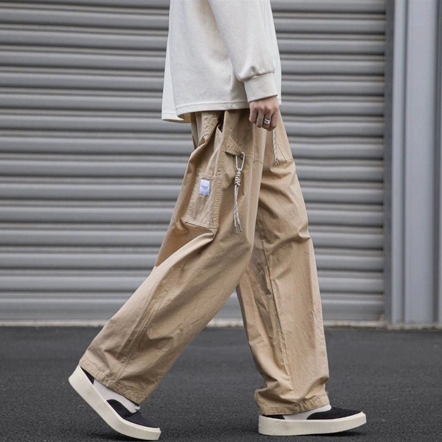 Khaki loose fit streetwear cargo pants shown from the left side with oversized pocket and drawstring detail, relaxed straight-leg silhouette, cream top, and casual shoes