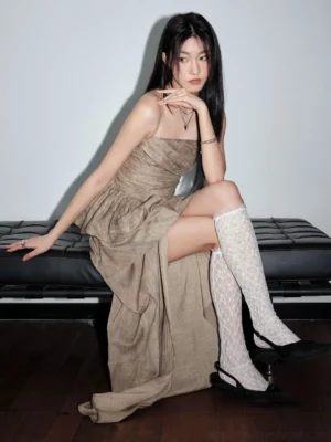 Three-quarter seated view of khaki ruched two-piece outfit styled with lace knee-high socks, black pointed heels, and layered necklaces