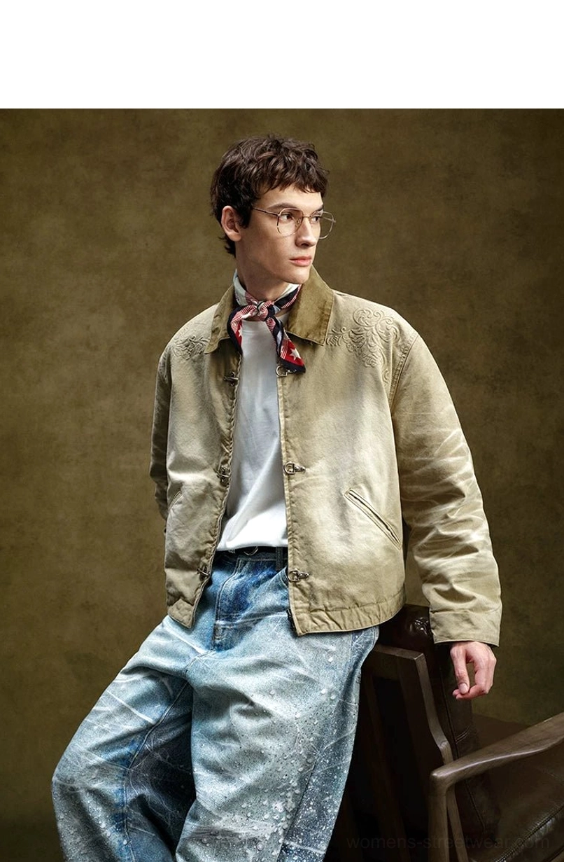 Male model wearing a relaxed cropped khaki toggle front utility jacket with floral shoulder embroidery, scarf, tee, and wide-leg jeans