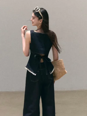 Back view of dark wash denim peplum cami top showing adjustable tie back and white lace trim with woven straw tote