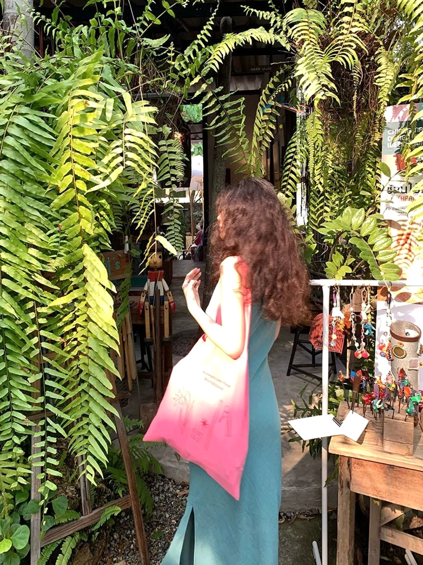 Back three-quarter view of lake blue sleeveless side slit maxi dress styled with a pink tote bag at an outdoor market