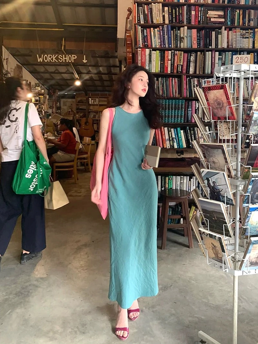 Lake blue sleeveless side slit maxi dress walking front view with pink tote bag in a retro bookstore setting