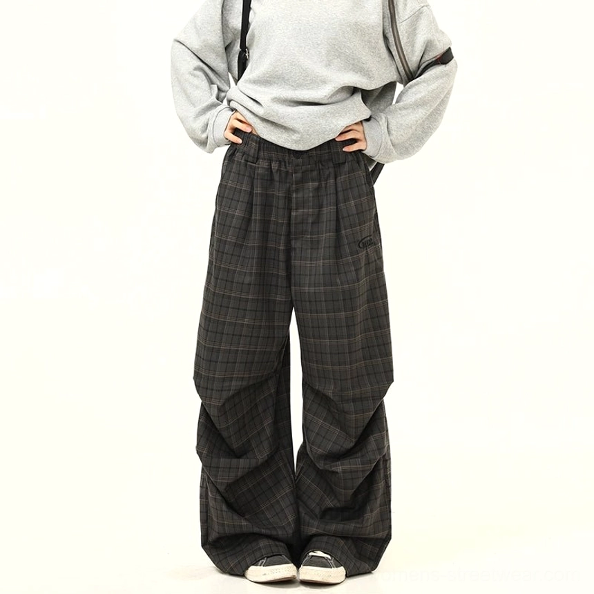 Low-angle front view of layered charcoal plaid wide-leg streetwear pants with loose draped legs and casual canvas sneakers