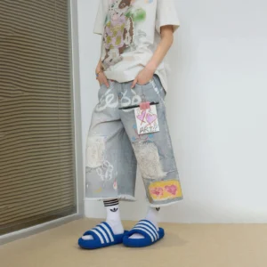 Light blue distressed graffiti denim Bermuda shorts shown from a low angle with wide-leg fit, raw frayed hem, patchwork details, heart artwork, graphic tee, socks, and blue slide sandals