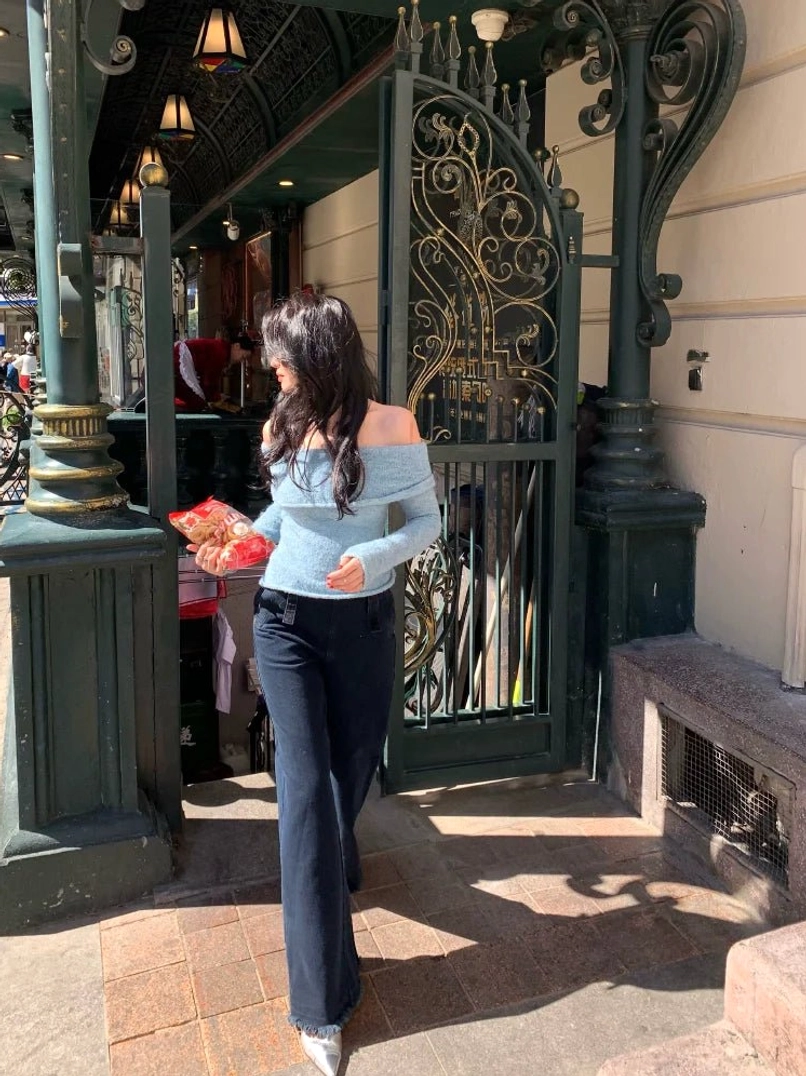 Light blue heathered off-shoulder knit sweater top shown from the back with dark straight-leg trousers in a city street outfit