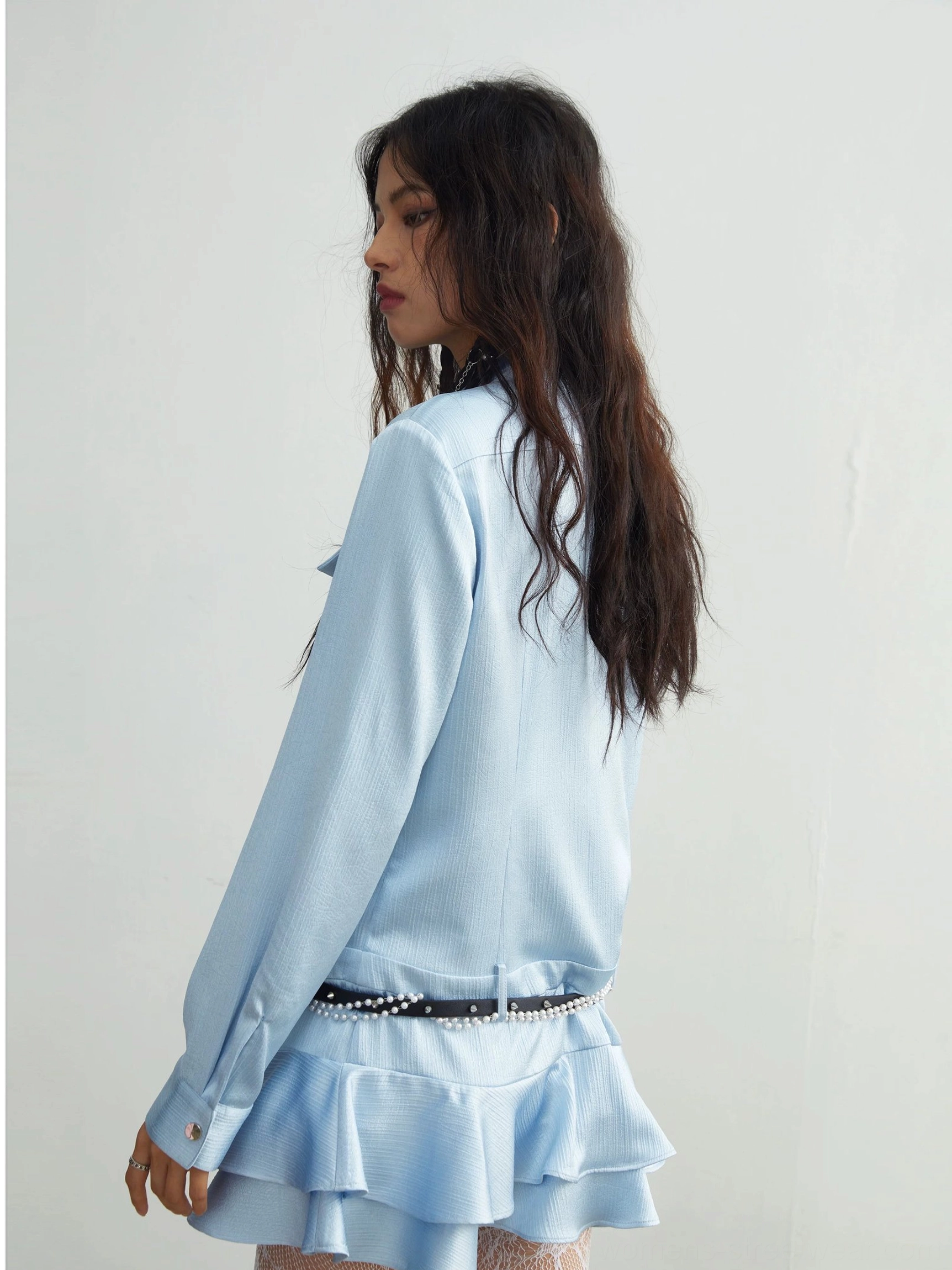 Back full-body view of light blue ruffle mini shirt dress with tiered hem, buttoned sleeve cuffs, and black pearl waist belt