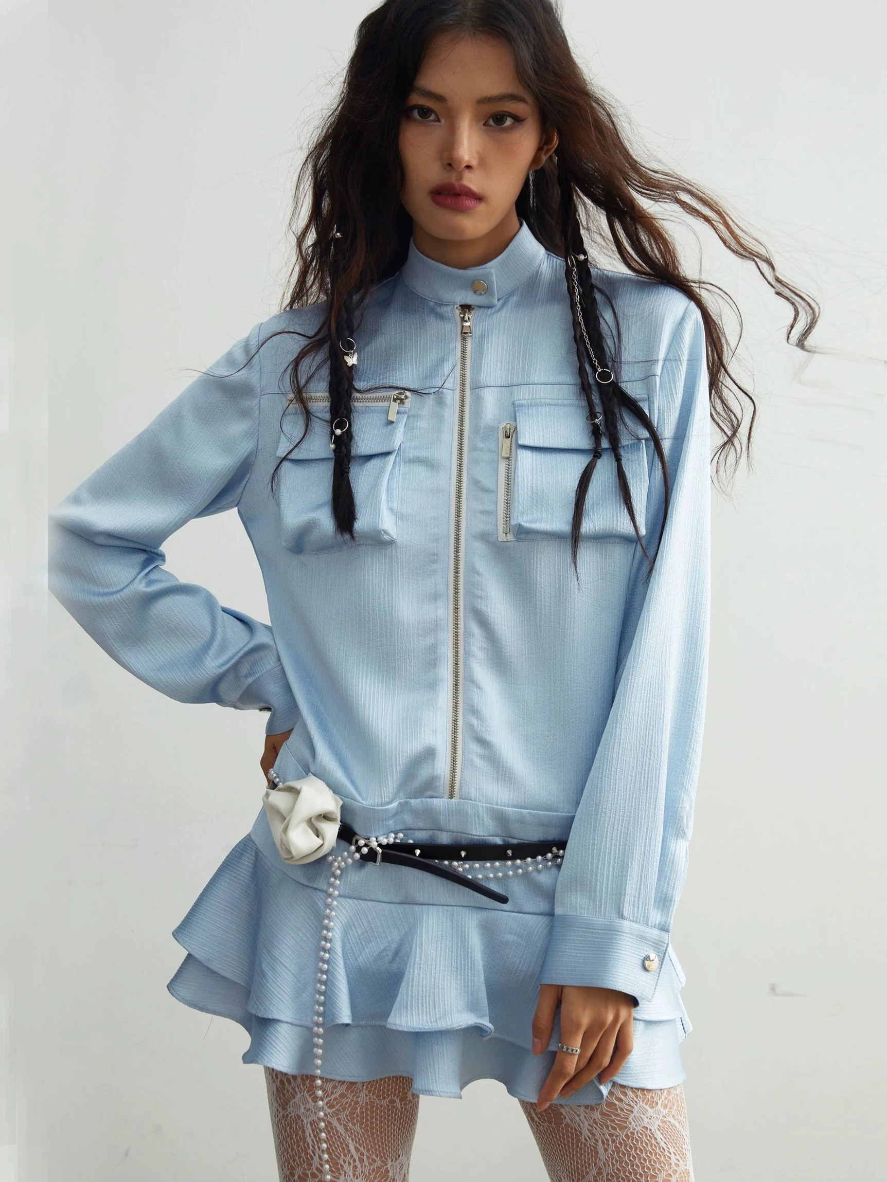 Front full-body view of a light blue utility ruffle mini dress with zip front, stand collar, chest pockets, pearl waist belt, and white flower accent