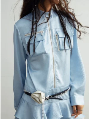 Close-up of light blue zip-front utility mini dress showing metal zipper, stand collar, dual chest pockets, ruffled waist, and pearl belt styling