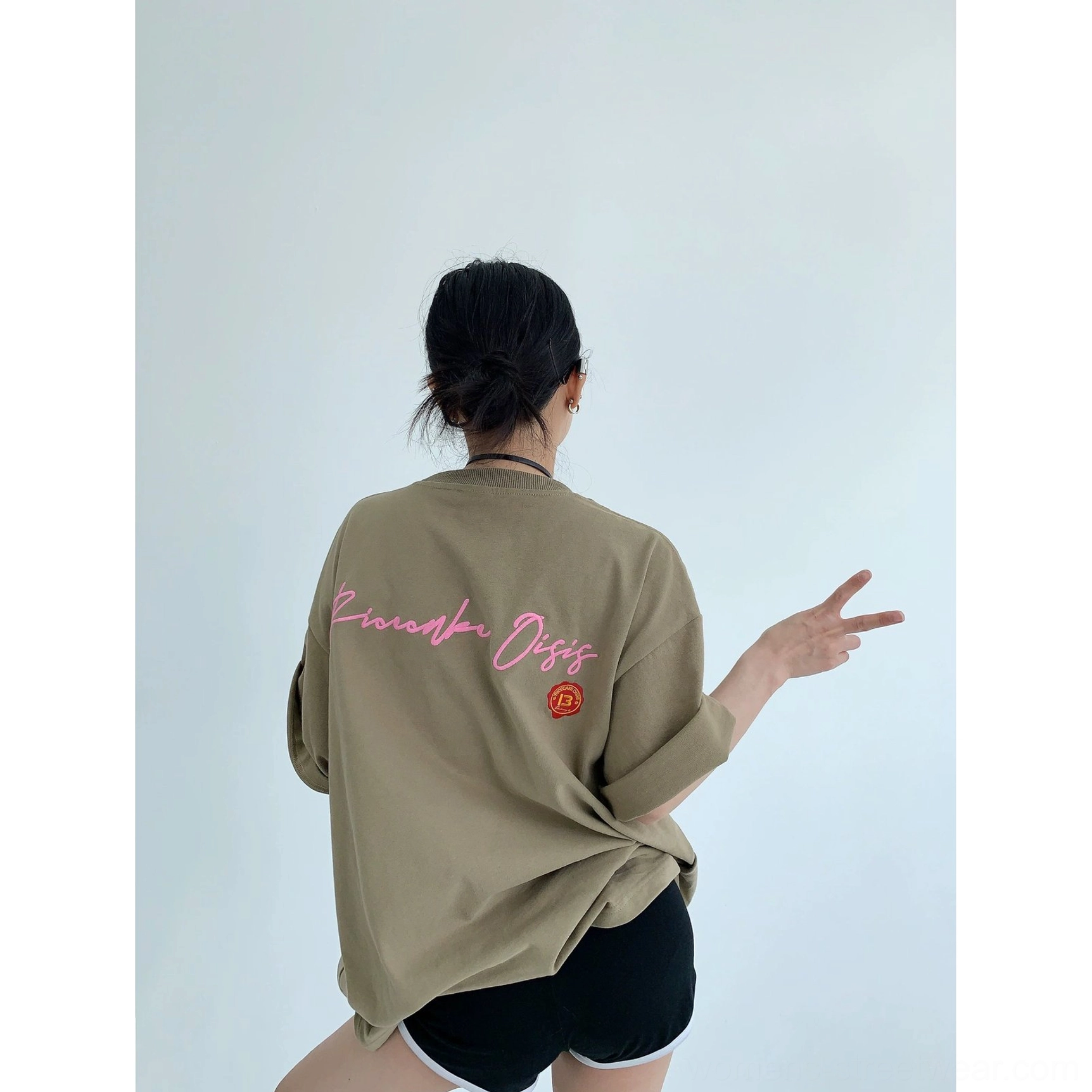 Light brown oversized streetwear T-shirt with neon pink cursive back graphic, orange emblem, relaxed fit, and casual shorts styling