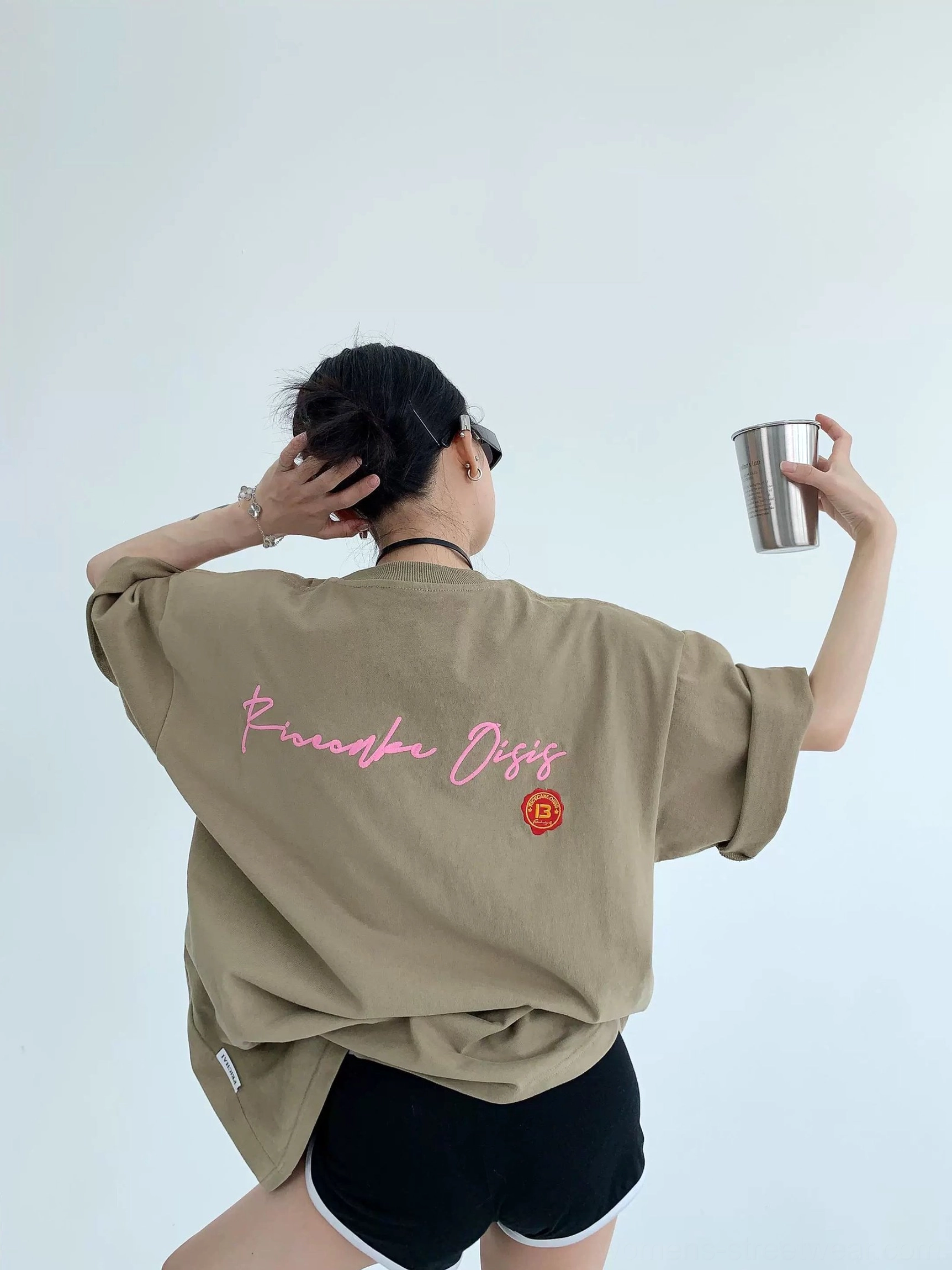 Light brown oversized graphic cotton tee with vivid pink cursive back lettering, orange circular logo, rolled sleeves, and black athletic shorts