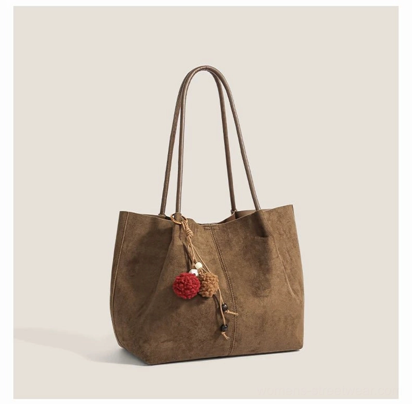 Light warm brown suede look tote bag shown from a front three quarter angle with dual shoulder straps and pom pom pendant