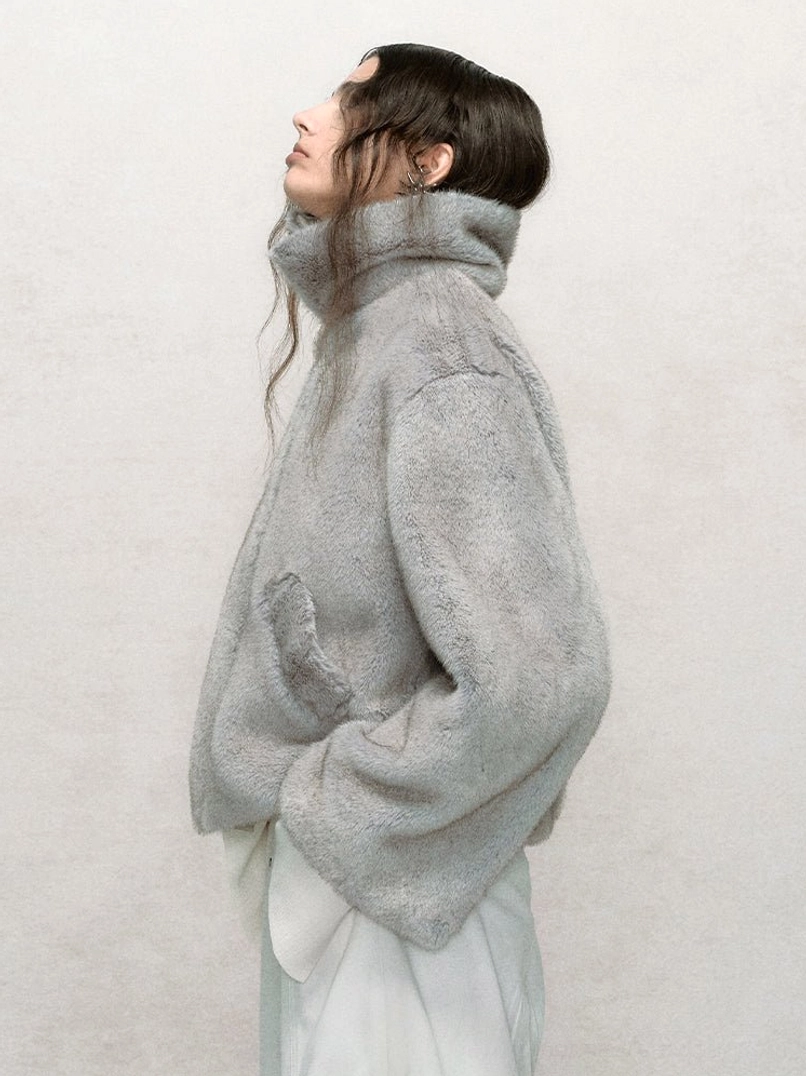 Light gray cropped faux fur bomber jacket shown from a side angle with high stand collar, fluffy fabric, relaxed sleeves, and short hem