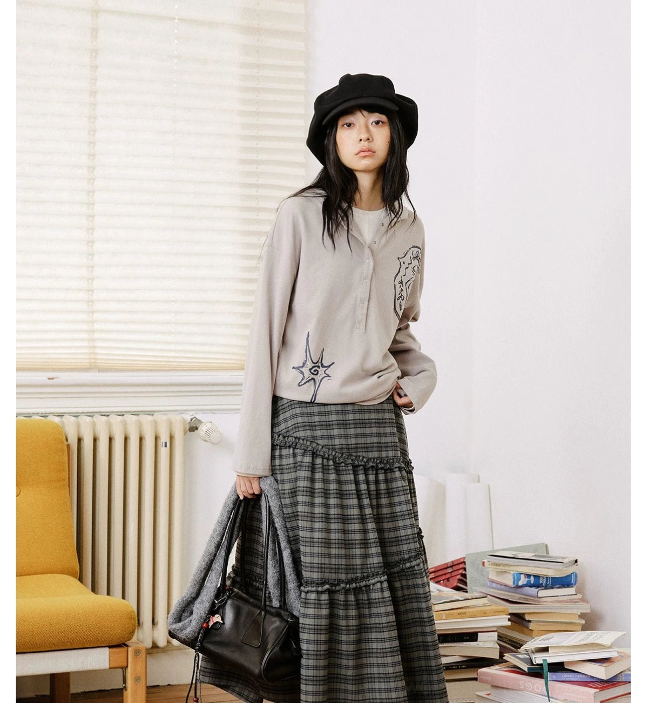 Light gray oversized embroidered graphic polo shirt paired with a plaid midi skirt and black leather tote bag