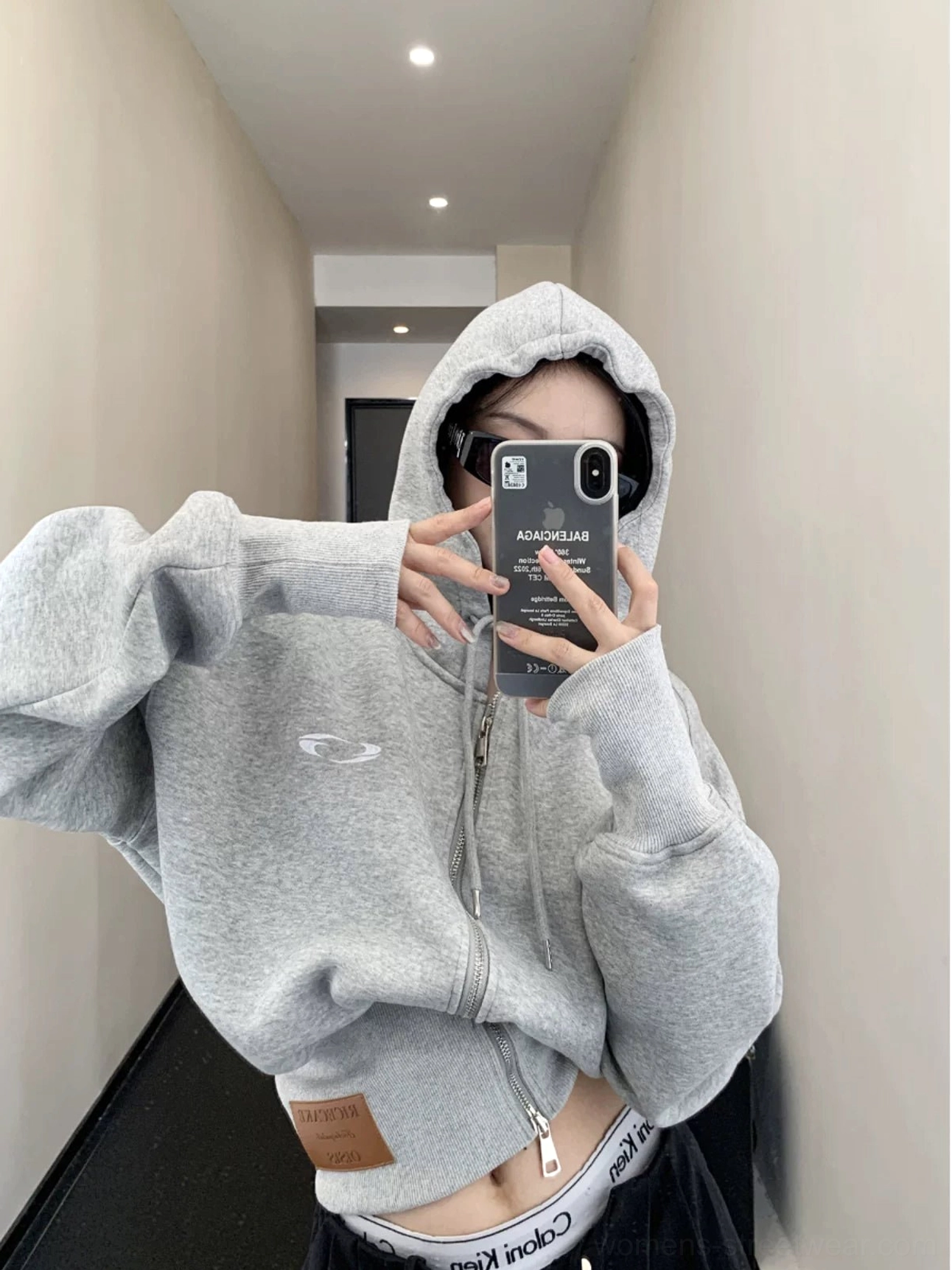 Front view of a light gray oversized cropped zip-up hoodie worn with the hood up, soft fleece texture, chest embroidery, and ribbed waist
