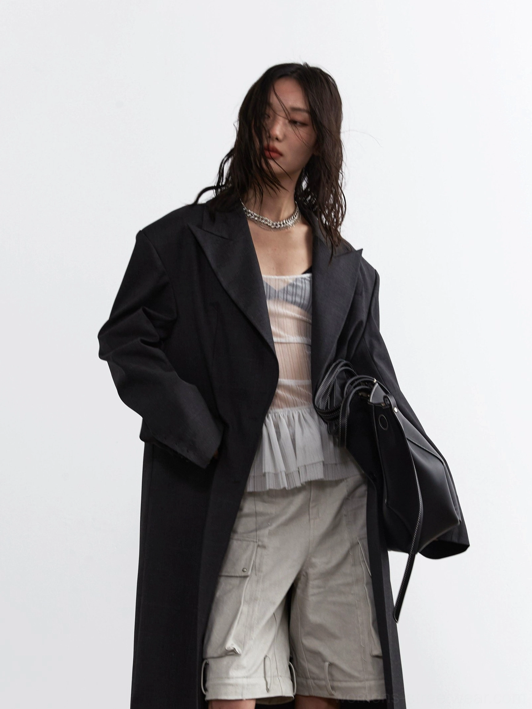 Side view of light gray sheer corset camisole with tulle peplum styled with black trench coat cargo shorts and leather bag
