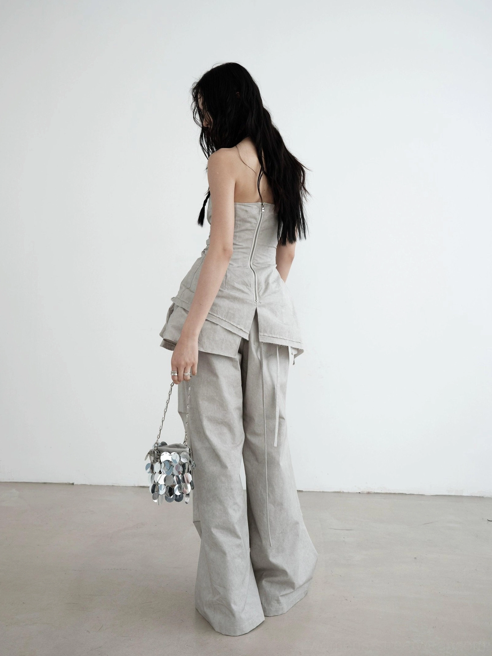 Back view of light gray strapless ruched asymmetric draped top styled with matching wide-leg utility pants