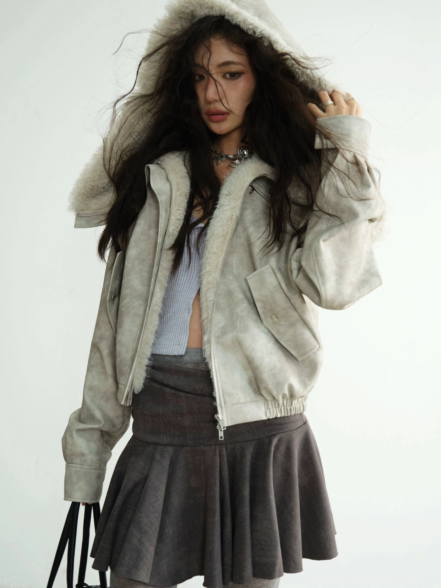Model wearing a light grey faux shearling hooded cropped jacket with plush fur hood and front zipper in a three-quarter studio view