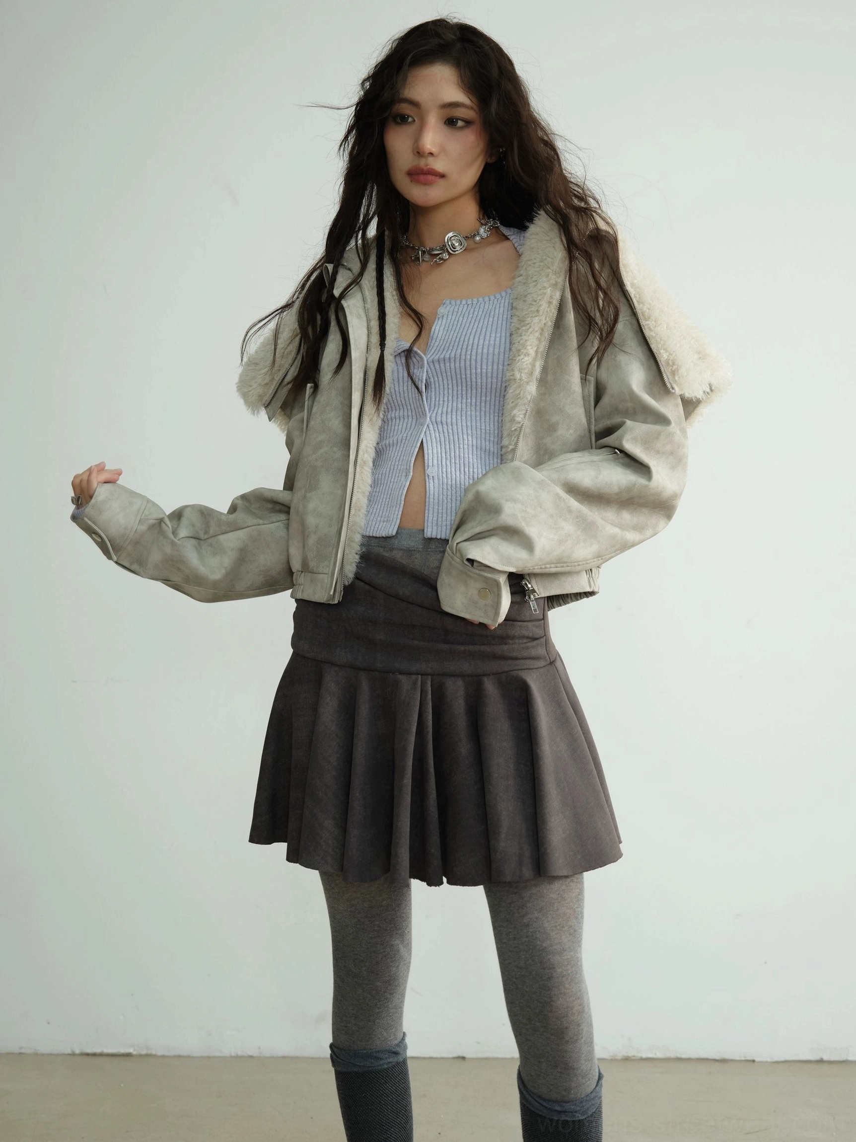 Light grey faux shearling hooded streetwear jacket worn open with cropped striped top and low-waisted pleated mini skirt