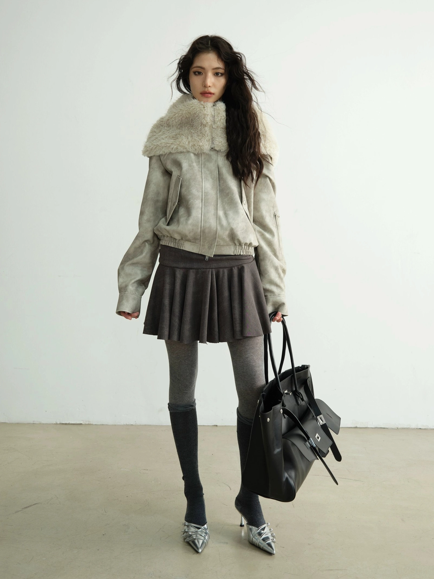 Full front view of an oversized light grey faux shearling hooded streetwear jacket styled with pleated mini skirt and thigh-high socks
