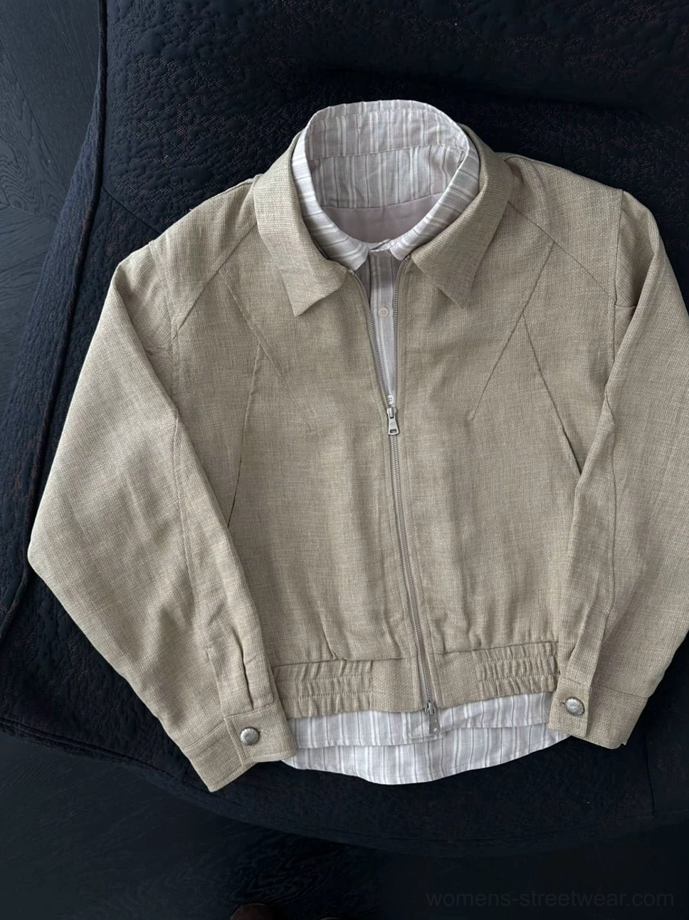 Top-down view of a fully zipped light khaki layered bomber jacket with striped collar and hem, gathered waist, and snap cuffs