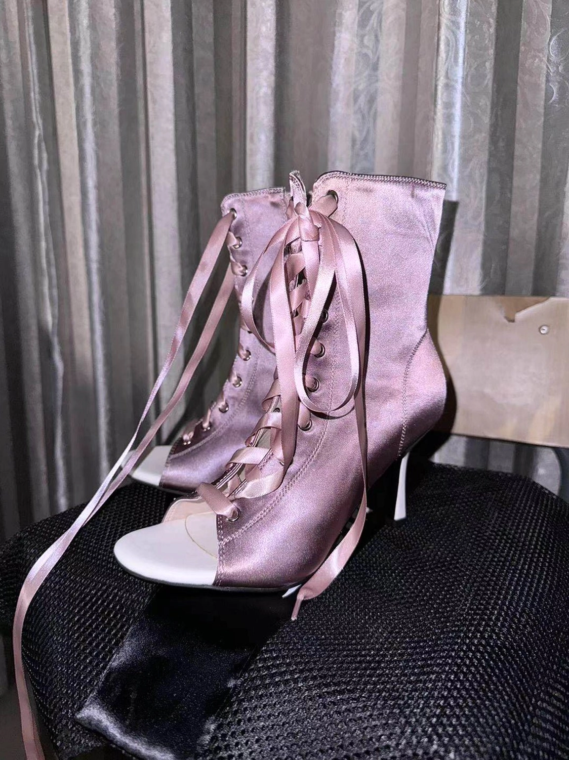 Light pink satin peep-toe ankle heels displayed on a black chair with long ribbon laces and hidden side zipper