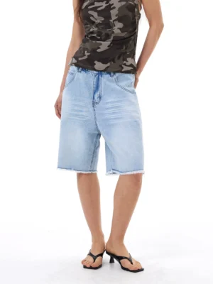 Full front view of light wash baggy denim Bermuda shorts with raw frayed hems styled with a camo tank top and black flip flops