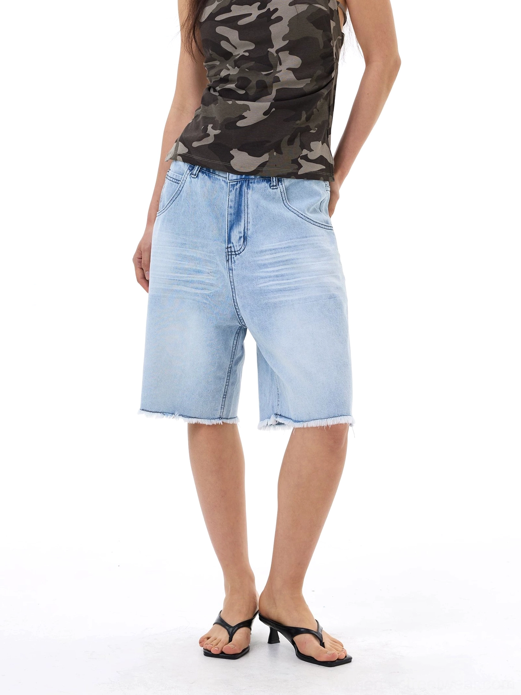 Full front view of light wash baggy denim Bermuda shorts with raw frayed hems styled with a camo tank top and black flip flops