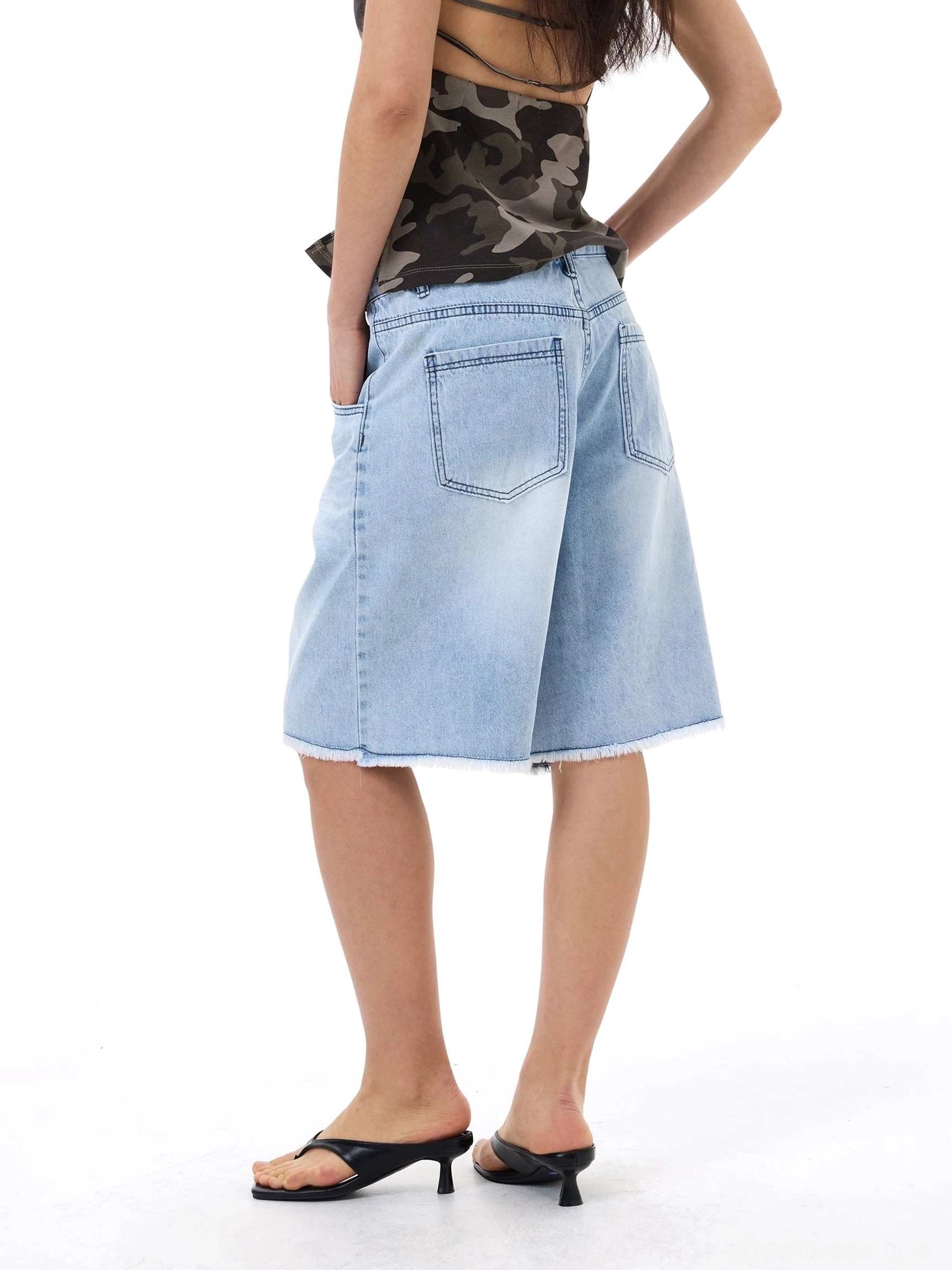 Rear side view of loose light wash denim Bermuda shorts showing back patch pockets and distressed frayed hems with heeled mules