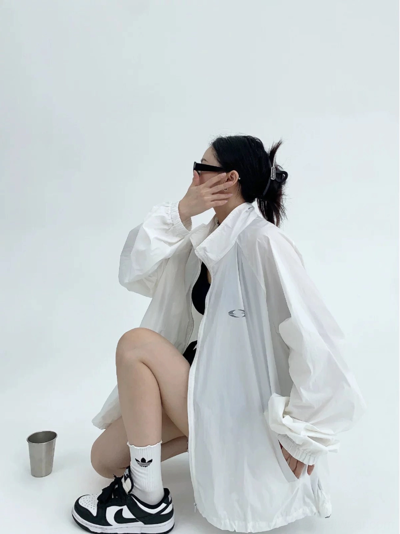 Side studio view of a woman wearing a lightweight oversized white windbreaker jacket with stand collar, subtle sheer fabric, small logo, and loose streetwear silhouette