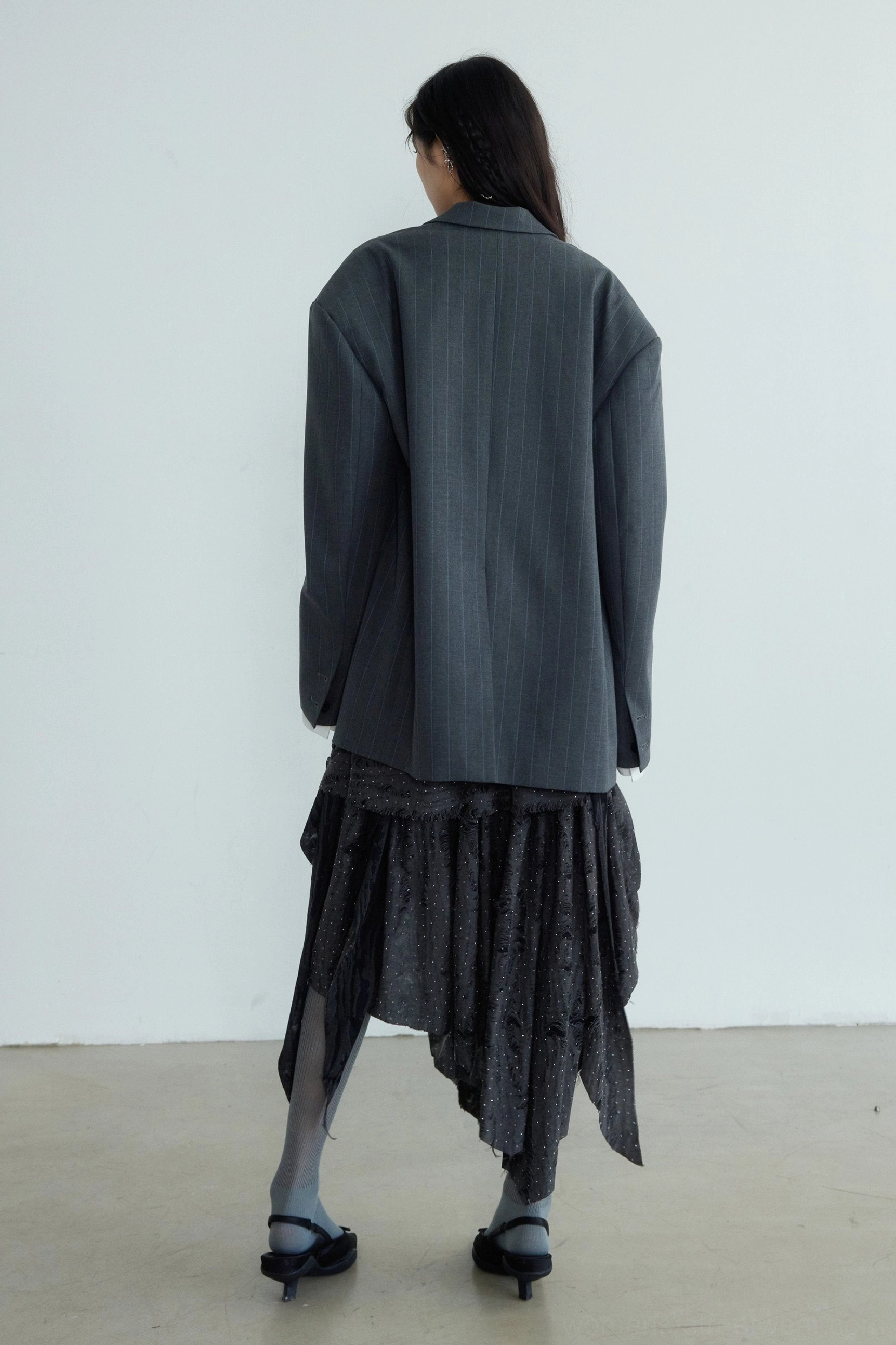 Complete back view of a long oversized gray striped blazer styled with an irregular ruffled skirt and low-heel shoes