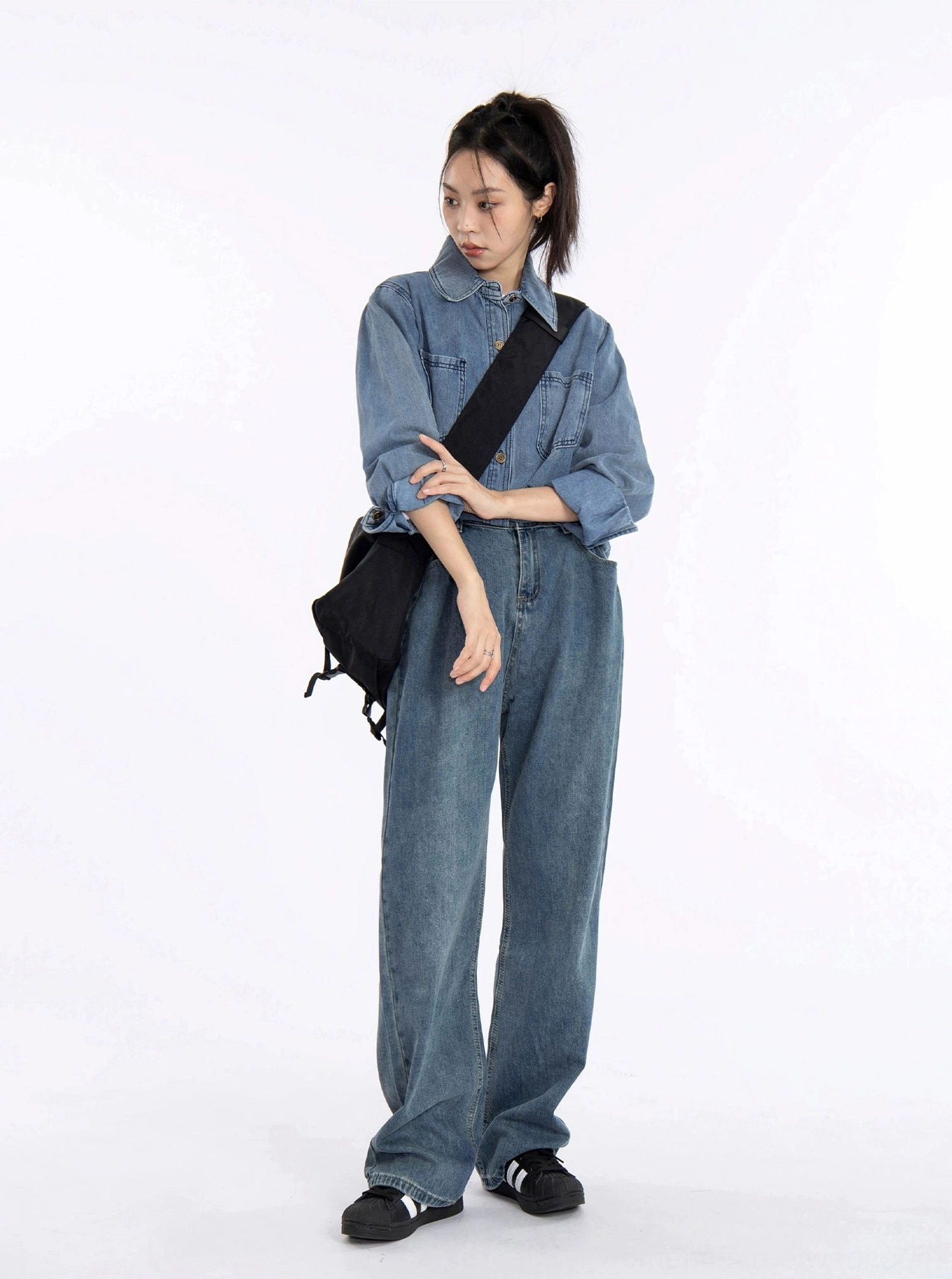 Three-quarter side view of a loose fit washed blue denim shirt jacket worn with matching denim pants and a black crossbody bag