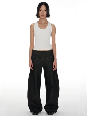 Model wearing dark indigo low-rise curved raw denim jeans with sculptural banana-leg wide silhouette and white tank top