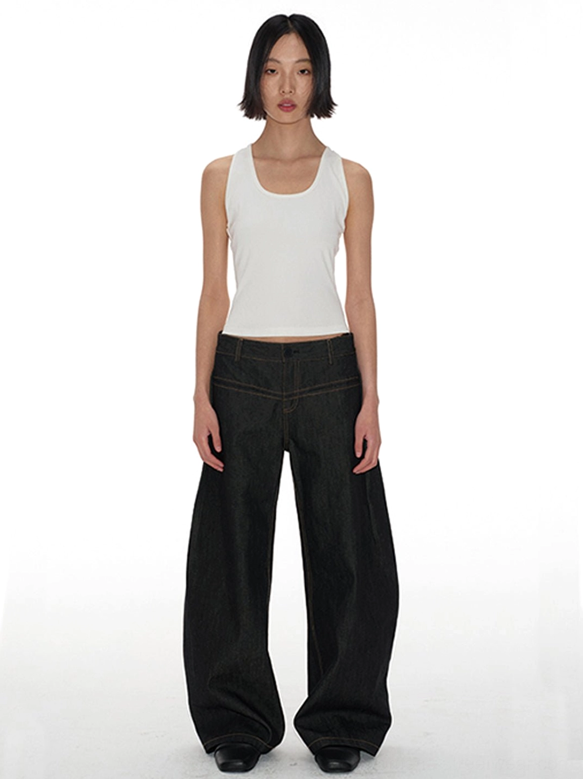 Model wearing dark indigo low-rise curved raw denim jeans with sculptural banana-leg wide silhouette and white tank top