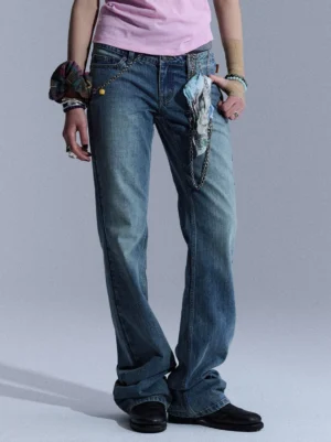 Front partial view of blue low-rise slim flare jeans with vintage wash, waist chain, and printed fabric accents styled with a pink cropped top