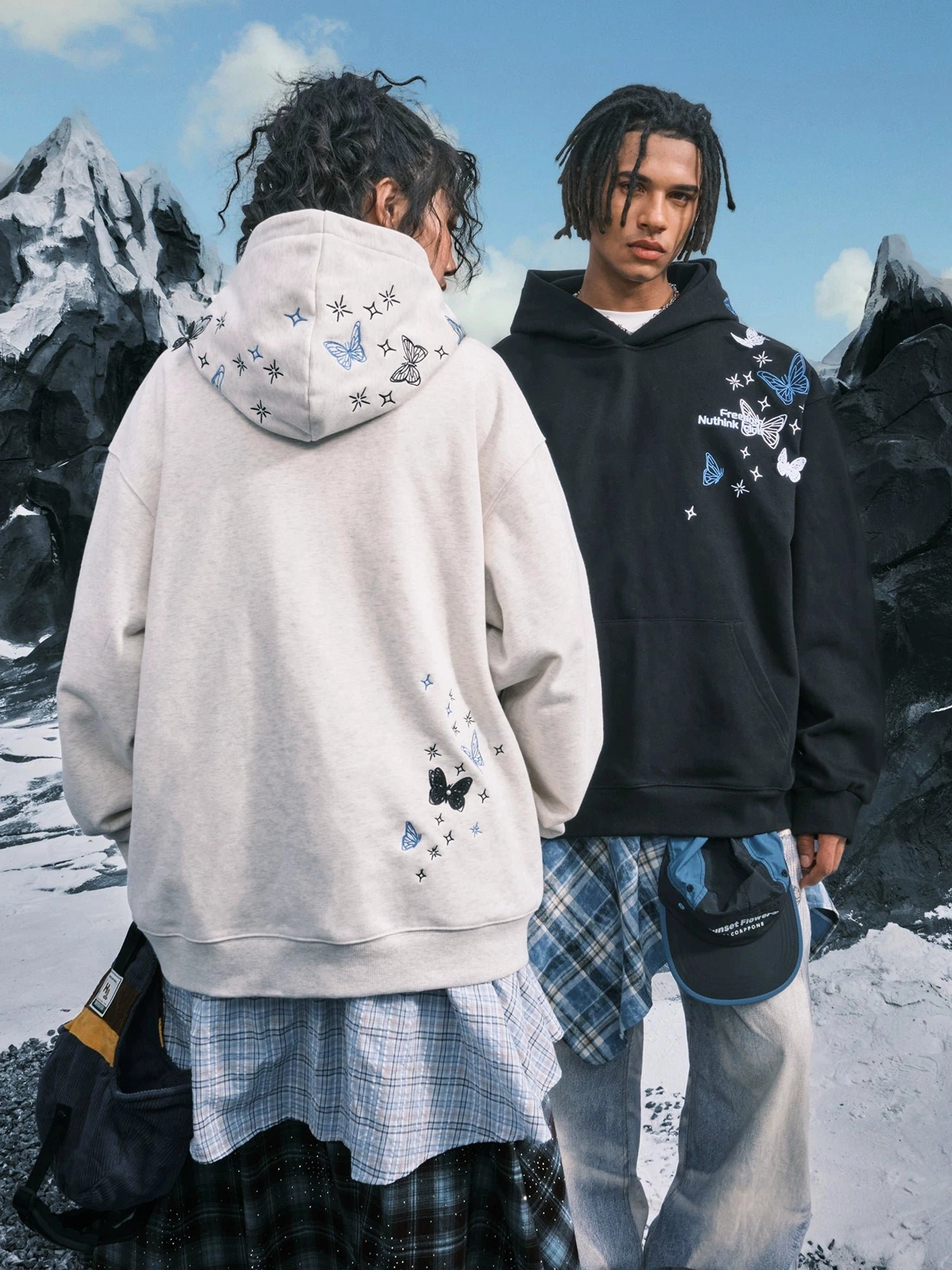 Rear view of a female model in a White Ash butterfly embroidered hoodie styled with a plaid skirt beside a model in a Black hoodie