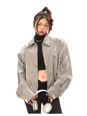 Metallic gray oversized faux leather streetwear jacket on a female model with dropped shoulders, snap button closure, embroidered chest badge, and black jeans