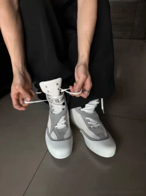 Person adjusting laces on white and grey metallic mesh high-top boxing boots worn with loose black streetwear trousers