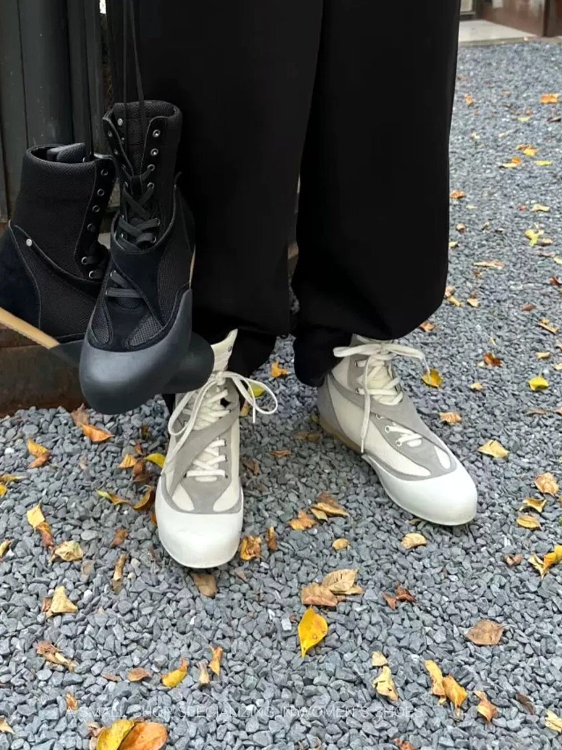 White grey mesh and black high-top boxing boots displayed on gravel with autumn leaves in an urban streetwear setting
