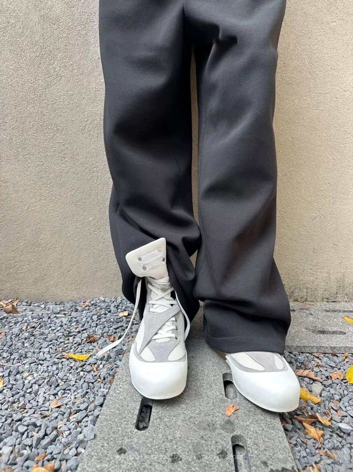 White and grey high-top boxing boots with low flat soles styled under dark wide-leg trousers on gravel pavement