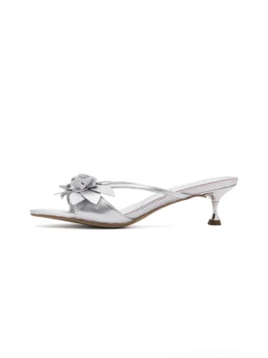 Metallic silver floral kitten heel slide sandals shown in a left side studio view with slim crisscross straps, dimensional flower embellishment, open toe, and tapered low heel